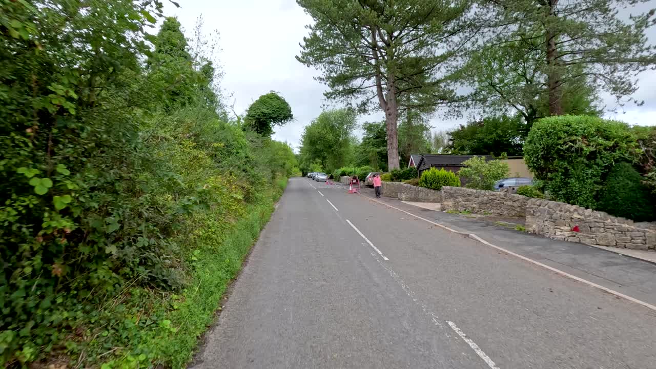 A vehicle travels down a quiet, tree-lined road in the English countryside, passing traffic cones and barriers under overcast daylight with steady forward camera movement