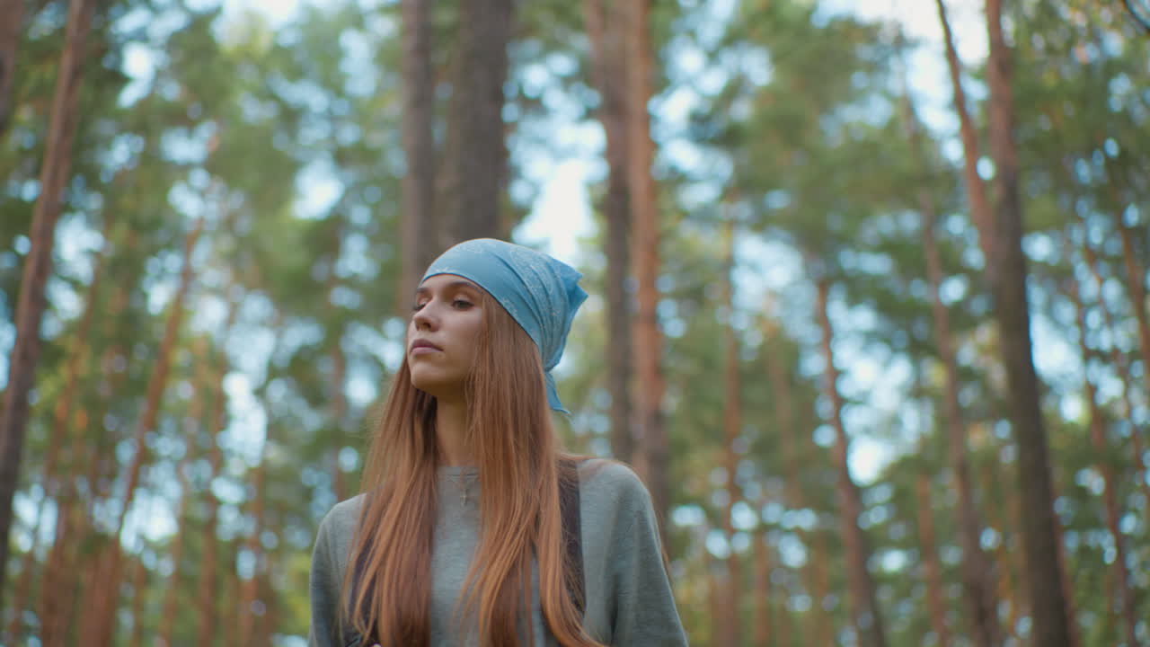 молодая женщина прогуливается по пышному лесу, глядя вверх с задумчивым выражением лица, в синей бандане и рюкзаке, в окружении высоких, ярких деревьев и естественной зелени