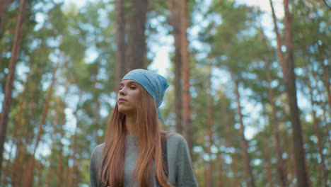 young woman hiking through lush forest, looking upward with contemplative expression, wearing blue bandana and backpack, surrounded by tall, vibrant trees and natural greenery