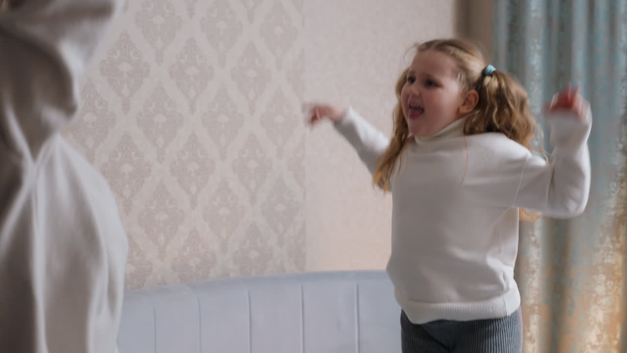 Mother and daughter in cozy sweaters joyfully dance together in living room, mother tossing hair with energy while daughter with pigtails sings and gestures with excitement, expressing carefree happiness
