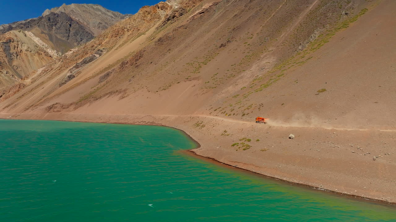 Orange mining truck on dirt road alongside turquoise lake, colorful Andes mountains, Chile. Aerial drone at low altitude, copy space
