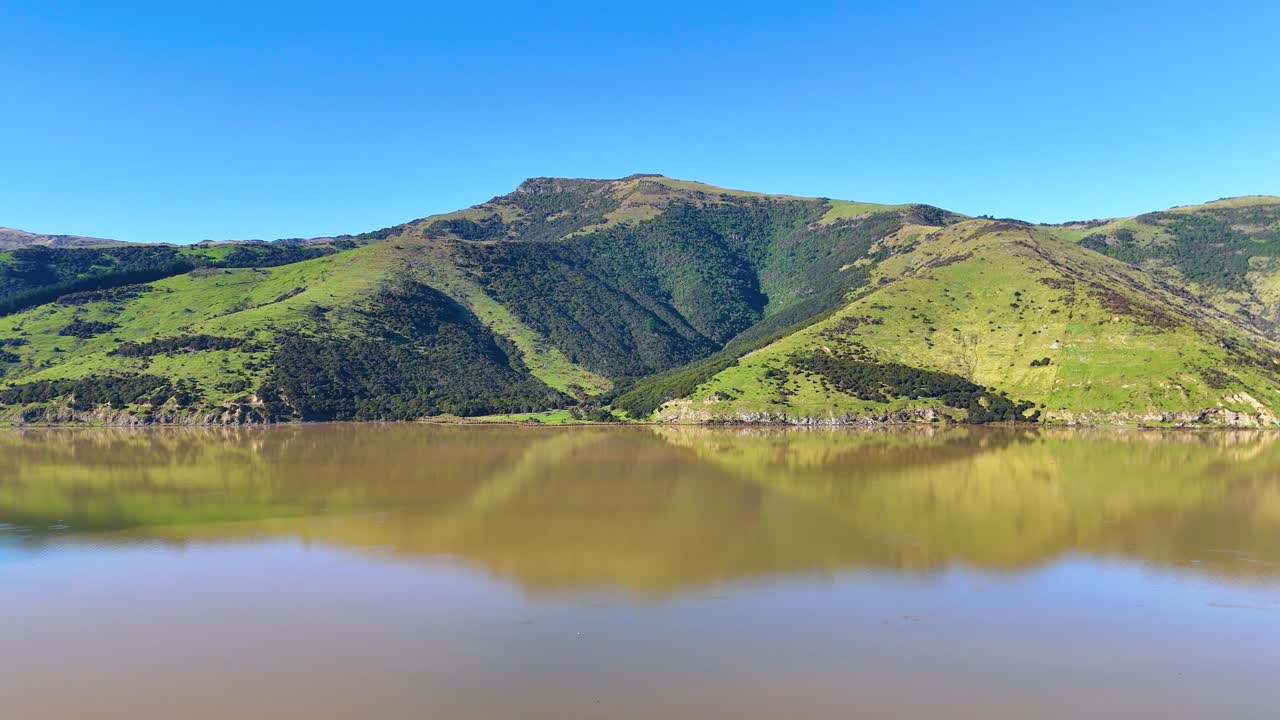 A tranquil scene of a lake reflecting lush green hills under a clear blue sky in Akaroa, New Zealand
