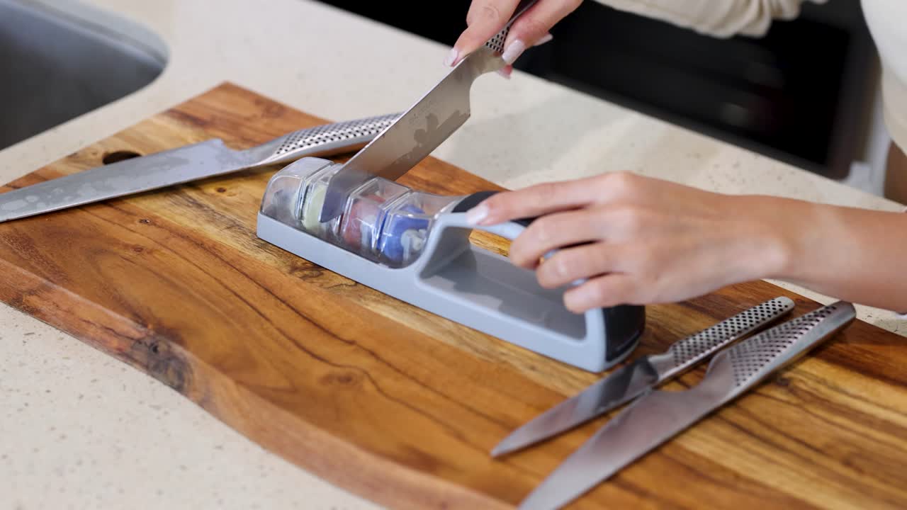 A person sharpens a knife using a sharpening tool on a wooden board in a well-lit kitchen