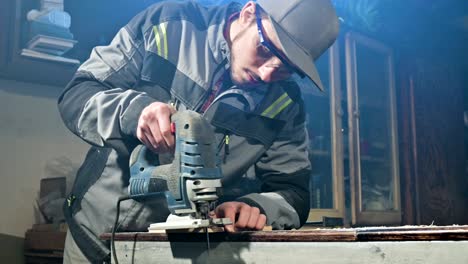 young man with a beard in gray overalls and goggles by profession a carpenter works as an electric jigsaw in his home workshop