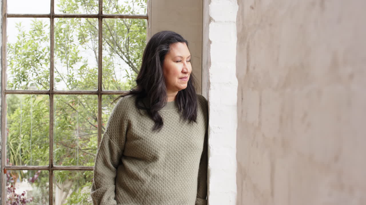 Thoughtful senior woman standing by window, enjoying peaceful moment at home