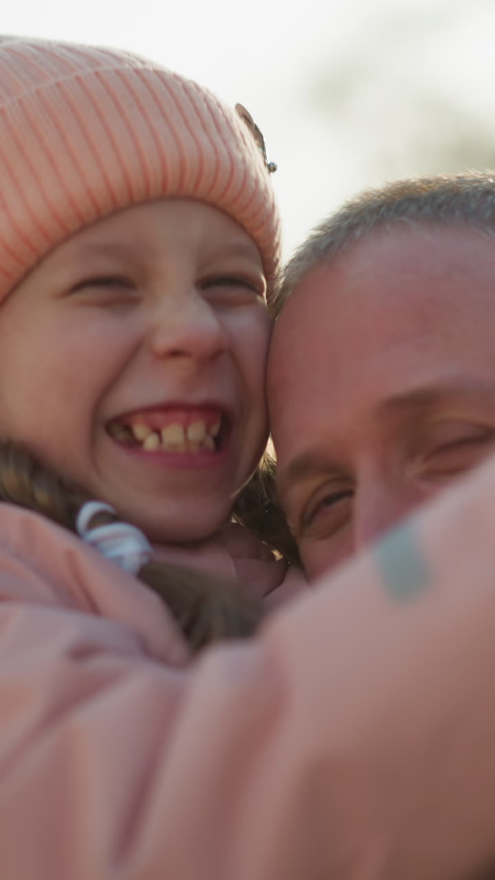 A man wearing a brown jacket carries a little girl in a pink cap and jacket as she makes a playful hand sign with one hand and places the other on his face. Both are smiling joyfully