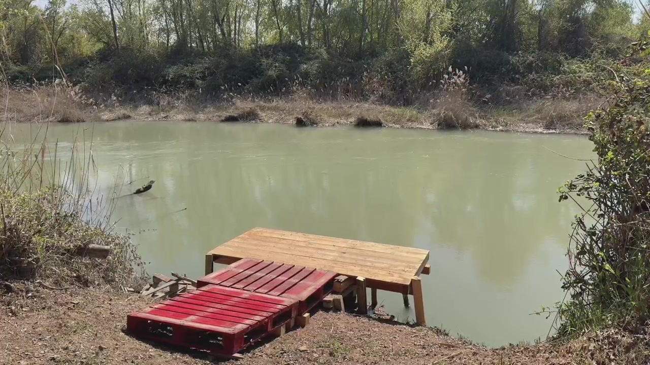A red-painted wooden fishing platform sits by the Tagus River as it flows through Aranjuez; riverside shrubs and trees frame the peaceful scene, while the empty platform awaits the anglers