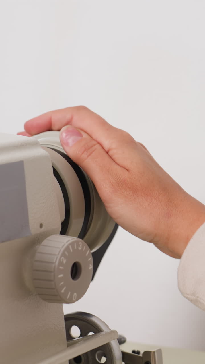 Nærbillede af hånd på symaskinehoved, skrædder justerer beige spændingsknap med et forsigtigt greb, hvidt ærme og metalkomponenter synlige, fokuseret studiebelysning, præcis bevægelse antyder håndværk, vedligeholdelse