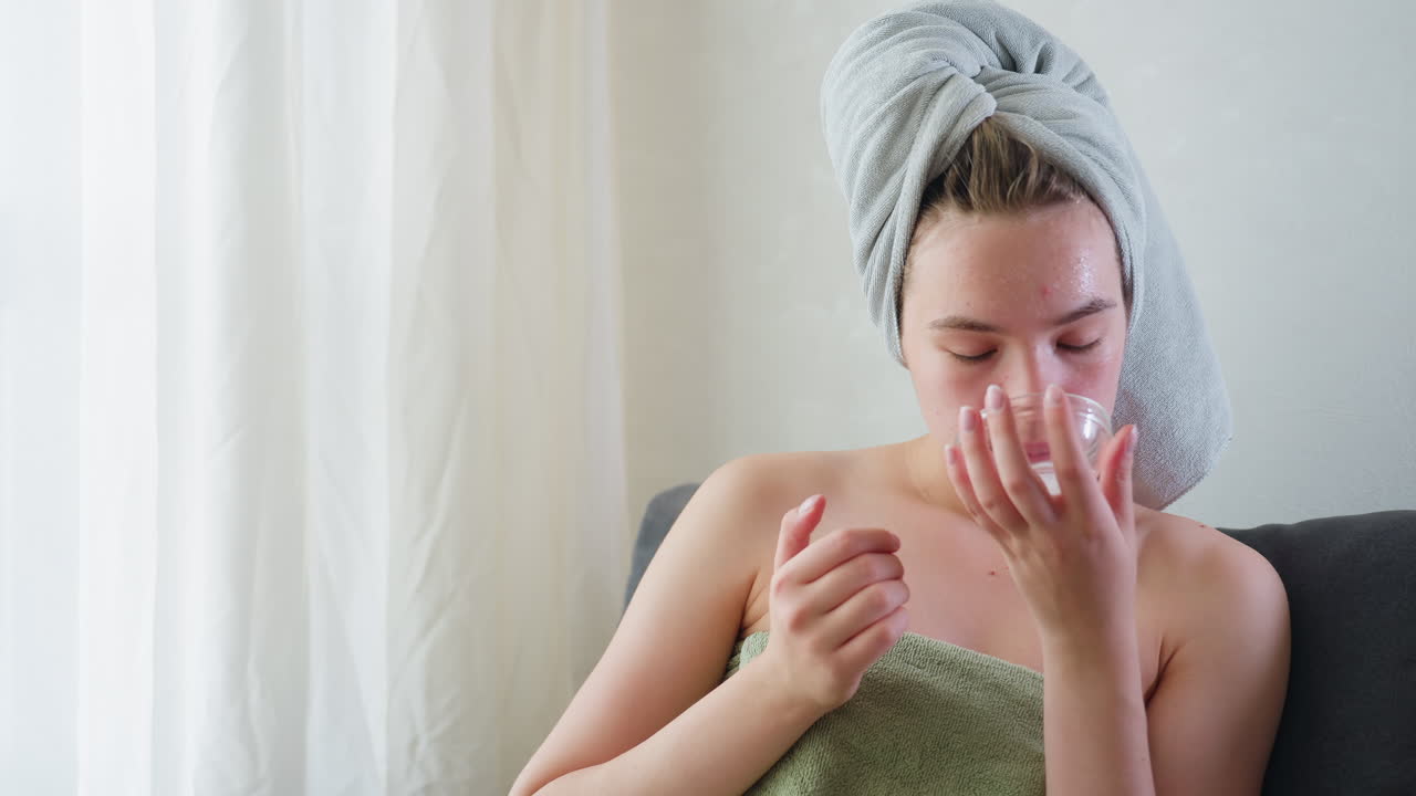 Young woman wrapped in towel with hair covered applies moisturizing skincare cream on face, sitting calmly indoors with eyes closed enjoying self care beauty routine in peaceful home setting