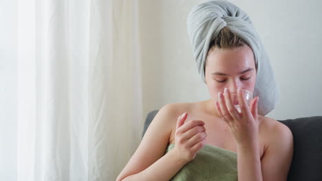 young woman wrapped in towel with hair covered applies moisturizing skincare cream on face, sitting calmly indoors with eyes closed enjoying self care beauty routine in peaceful home setting
