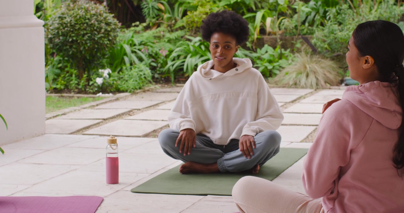 Ett mångfaldigt kvinnligt par pausar efter yoga på yogamattor på uteplatsen. En kvinna i rosa huvtröja tar en flaska, den andra i vit huvtröja ler.