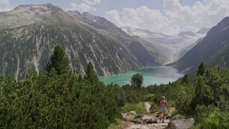 A-caucasian-girl-in-a-white-dress-walks-down-a-path-towards-a-blue-lake-and-a-beautiful-mountain-scenery