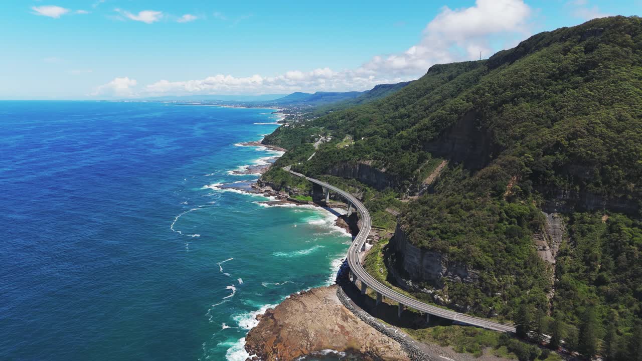 Driving cars on curvy coastal road in Australia. Famous Sea Cliff Bridge on Lawrence Hargrave Drive. Sunny day with blue sky and Blue ocean. Aerial backwards wide shot.