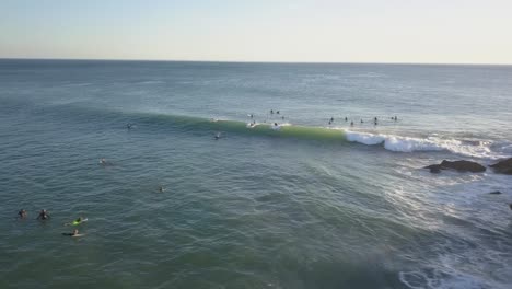 Panorámica-Aérea-De-Un-Gran-Grupo-De-Surfistas-Flotando-En-El-Agua-Mientras-Algunos-De-Ellos-Atrapan-Olas-Que-Pasan,-Con-Agua-Blanca-Rompiendo-Y-Mar-Azul-verde---Portugal