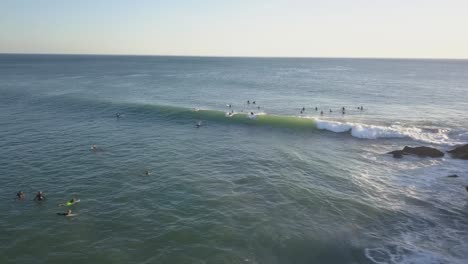 una panorámica aérea de un gran grupo de surfistas flotando en el agua mientras unos pocos de ellos atrapan las olas que pasan, con el agua blanca que se estrella y el mar azul-verde - portugal