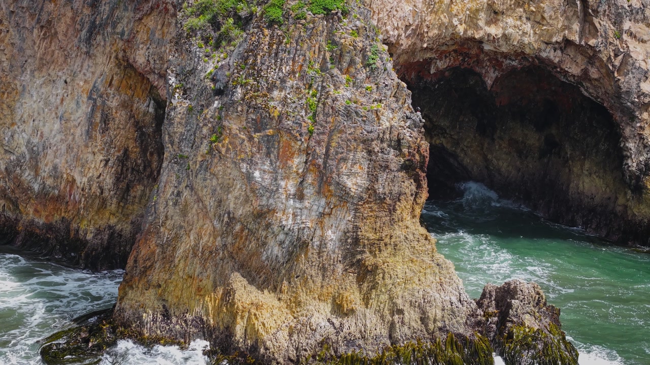 Aerial drone view of a grass-covered sea stack with a sea cave on Chiloé Island, Patagonia’s Pacific coast, Chile. Powerful waves hit a remote sandy beach and rugged green cliffs under an overcast sky