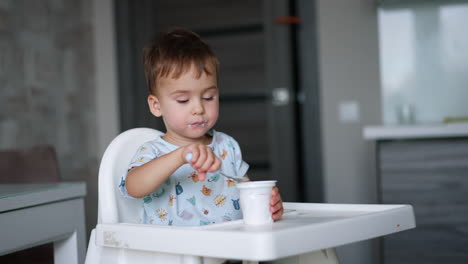 cute male kid enjoying yogurt. toddler sits in the kitchen eating dairy from a plastic cup. blurred backdrop.