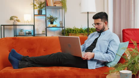 Indian-man-typing-working-on-laptop-using-apps-browsing-internet-lying-study-education-on-home-sofa