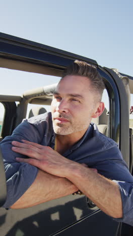 vertical video of happy caucasian man in car at beach, leaning out of window enjoying the view