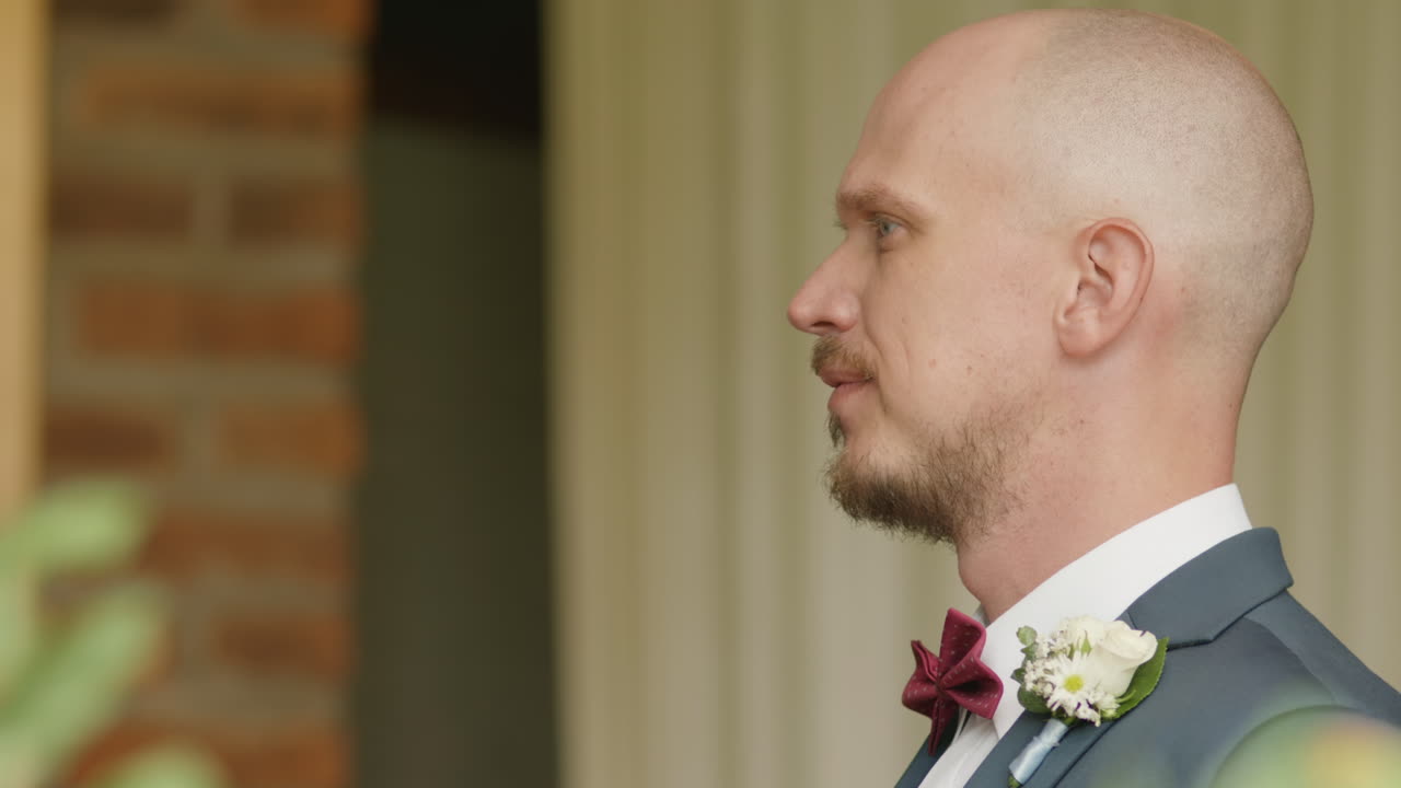 Medium shot of a bald groom with blue eyes, a mustache, a smart suit, a bow tie, and a flower in his lapel. He's smiling with a cheerful expression.