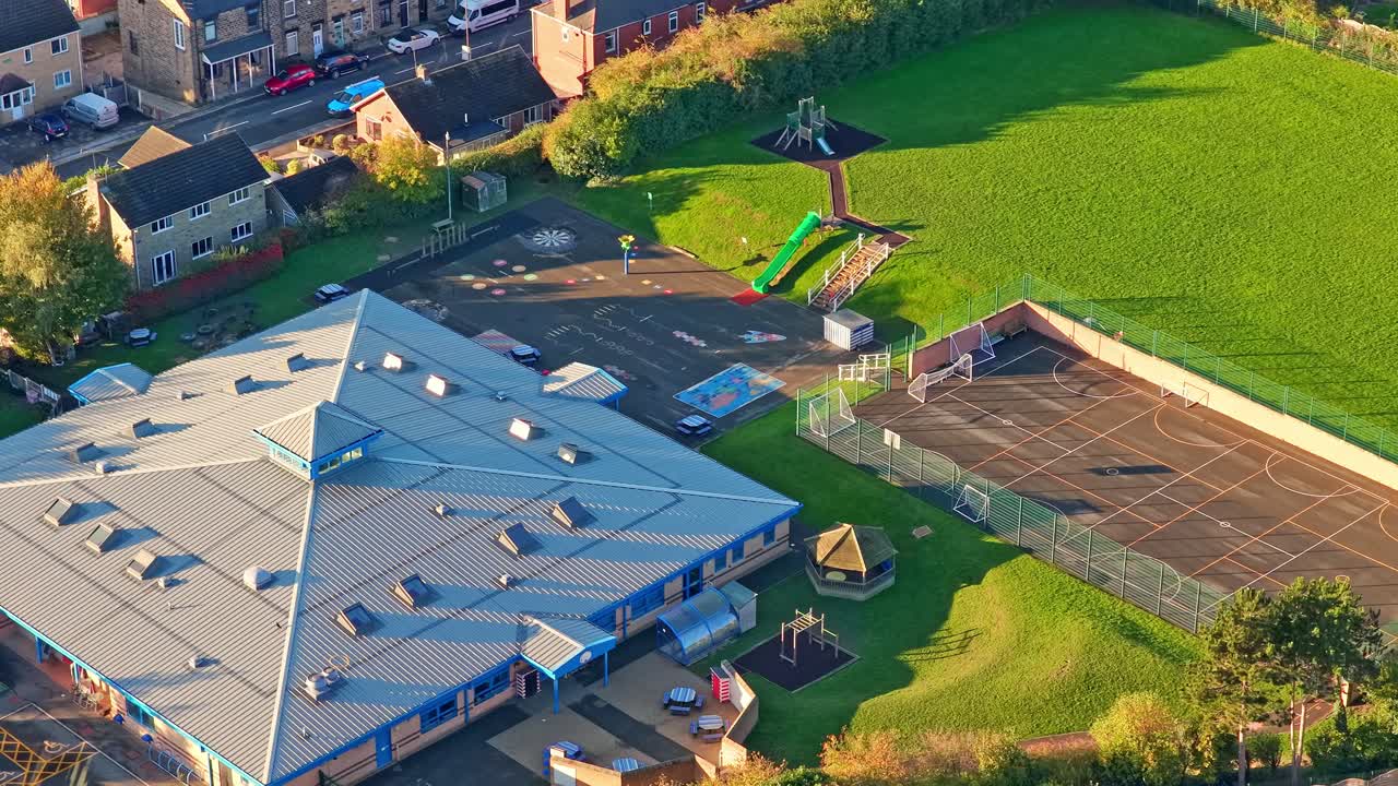 Hemingfield Primary School’s hexagonal roof, colorful play equipment, tennis court, and grassy field are visible in soft morning light during an ascending aerial over South Yorkshire