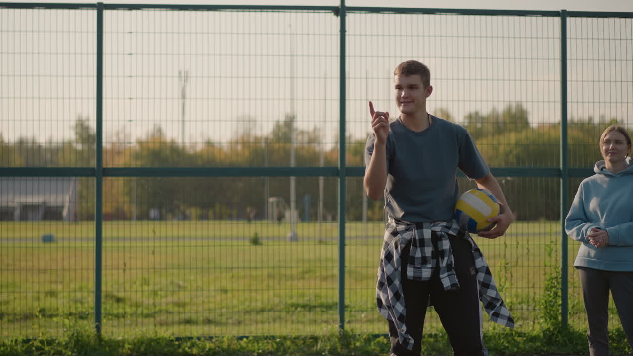 instructor de voleibol con la pelota bajo el brazo apuntando al jugador en el fondo, con la dama con capucha mirando, instalación deportiva en el fondo con entorno al aire libre activo