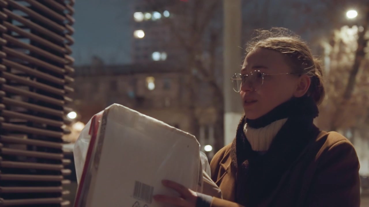 Donna bianca con una scatola da asporto di notte vicino a una recinzione metallica e un lampione, avvolta in un cappotto marrone e una sciarpa, mani guantate che maneggiano un contenitore di cartone, scena con tastiera e armadietto, pioggerella serale e atmosfera cinematografica.