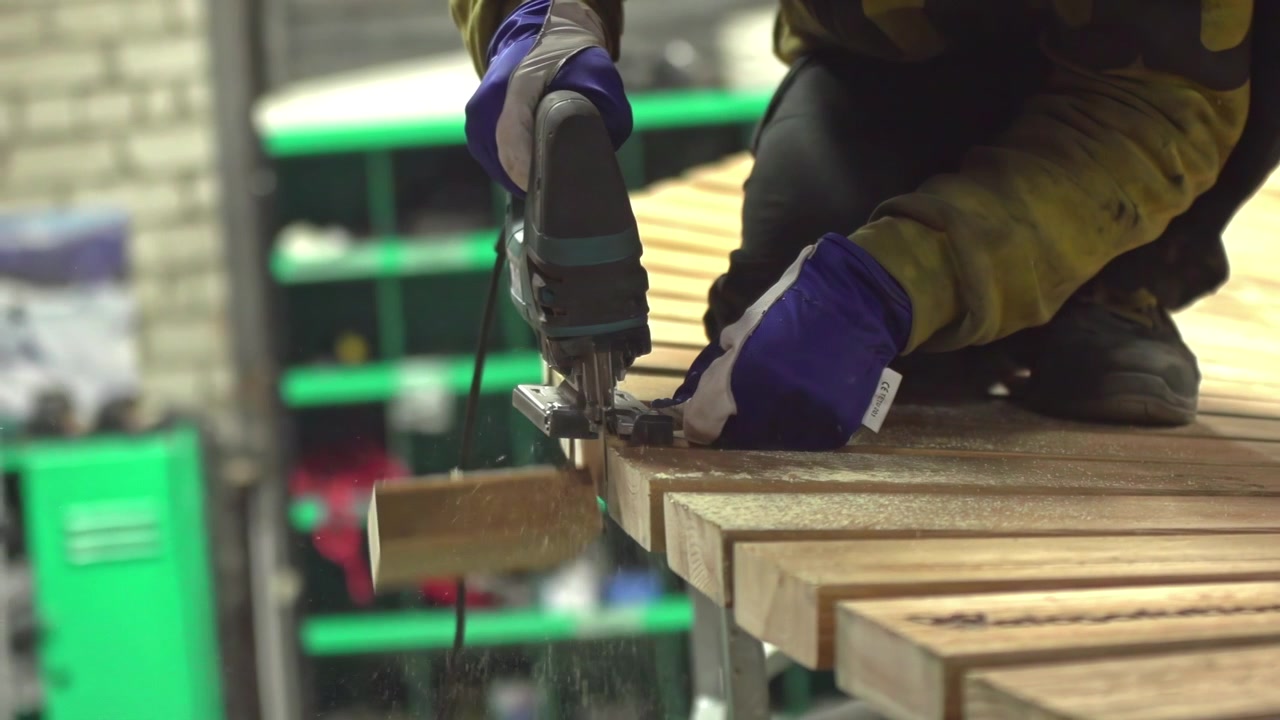Carpenter Is Cutting A Wooden Plank With Jig Saw Machine Using Gloves. Man Sawing A Wood Plank With Electric Jig Saw Machine In Carpentry