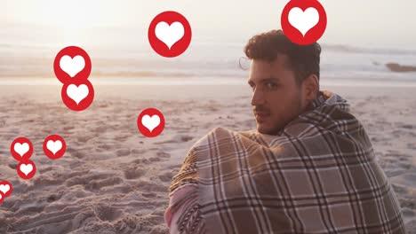 Multiple-heart-icons-floating-against-portrait-of-caucasian-man-with-blanket-smiling-on-the-beach