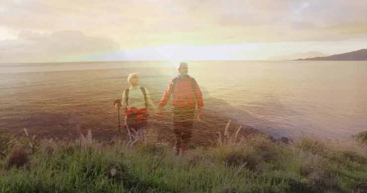 Sol a brilhar na água, impulsionando um casal de idosos a fazer trilha, fincando o bastão, caminhando pela duna em direção ao pôr do sol.