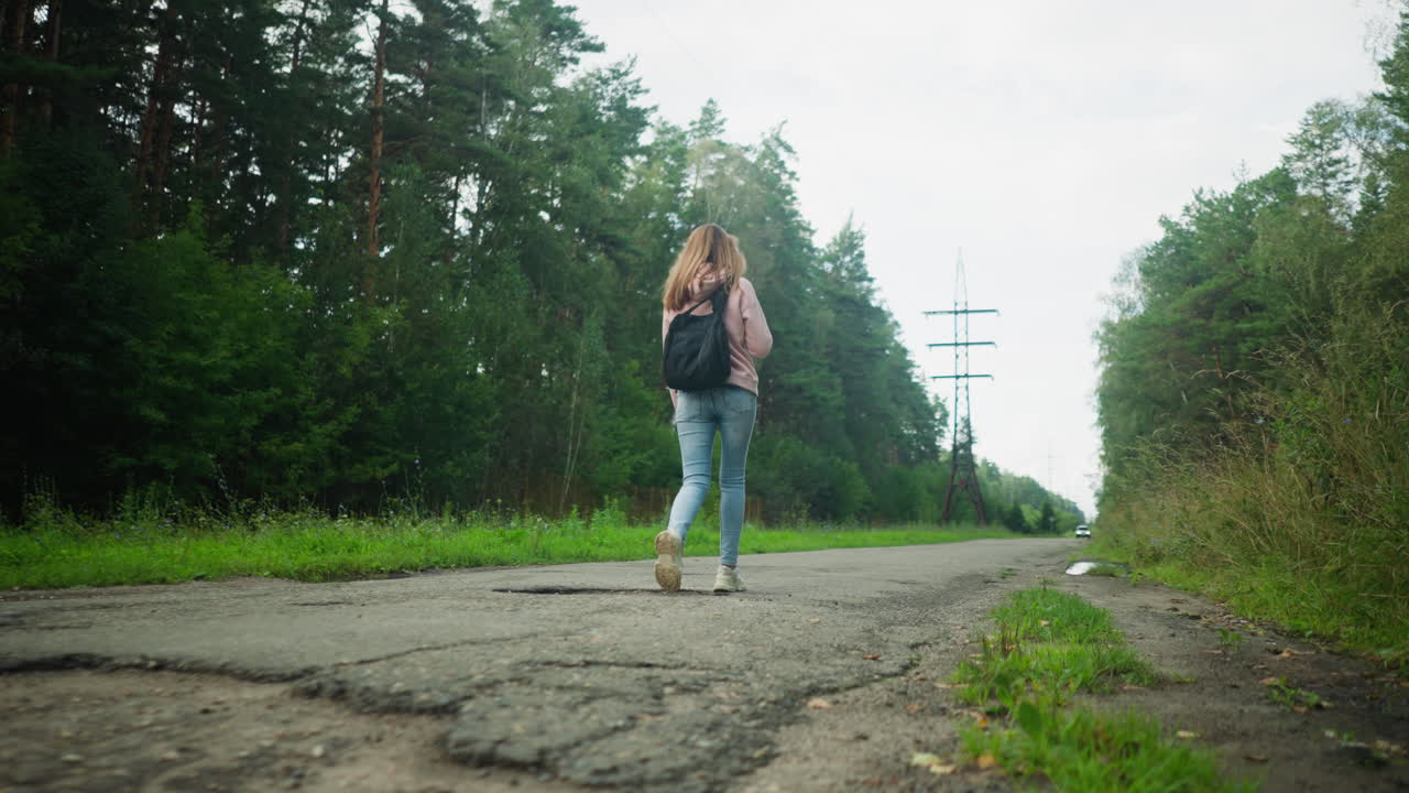 Rear view of tourist walking alone on cracked rural road surrounded by dense forest and tall grass, carrying black backpack on shoulders, with power poles in distance