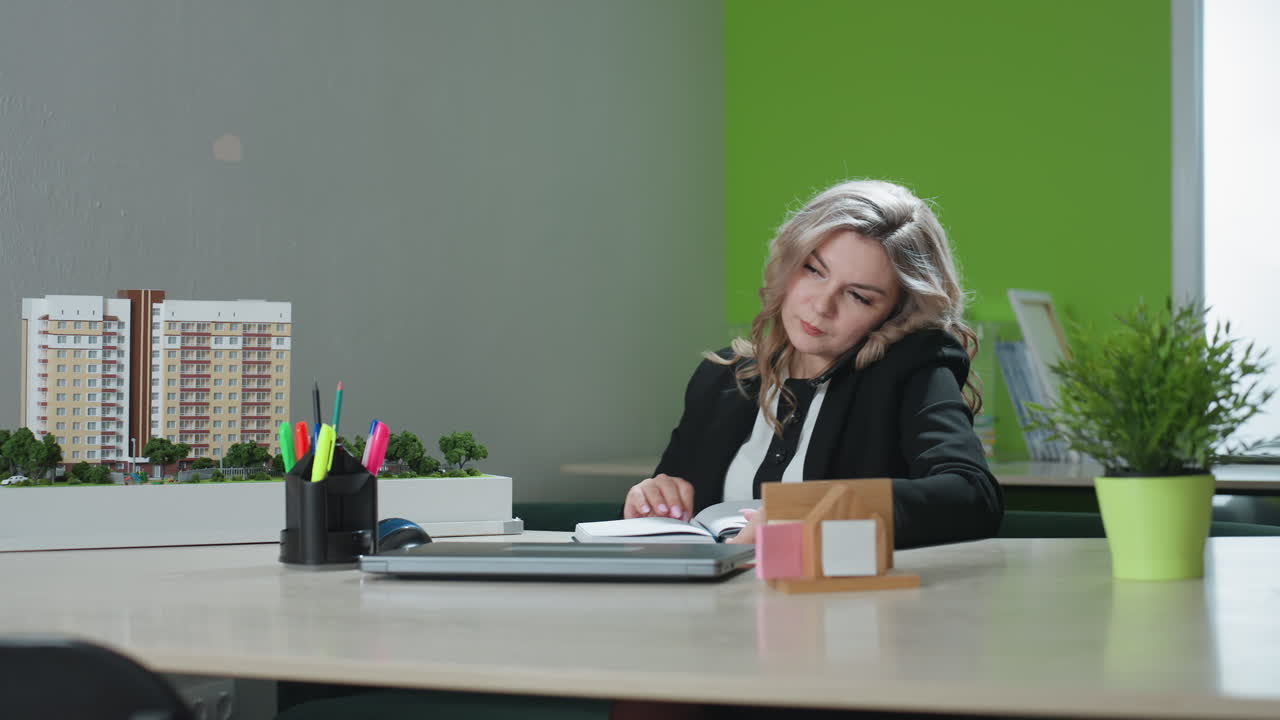 Real estate agent answers client call while multitasking at desk, opening book and preparing to take notes with pen, surrounded by architectural model and office supplies in modern workspace