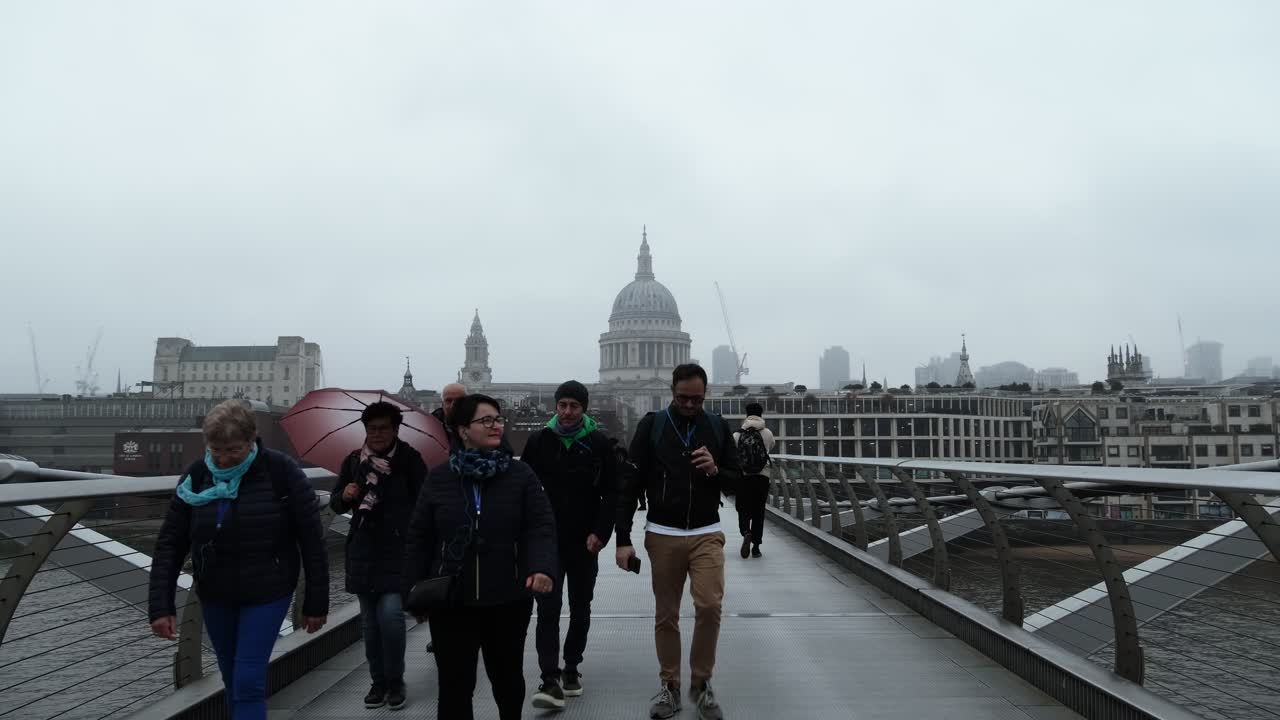 A scenic view of pedestrians walking across the Millennium Bridge towards St. Paul's Cathedral in London, during a misty rainy day.
