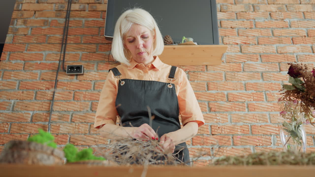 Woman in black apron working with dry twigs on wooden table against brick wall, focusing on natural craft preparation with decorative moss creating handmade rustic arrangement in cozy workshop environment