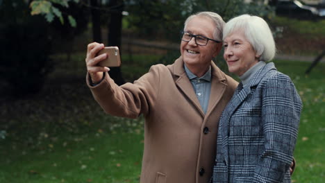 elderly couple posing and smiling to the camera while taking selfie photo with smartphone in the park at sunset 1