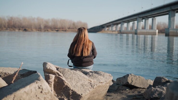 Donna seduta su grandi rocce vicino a uno specchio d'acqua calmo, di spalle alla fotocamera, rivolta verso alberi lontani e un cavalcavia. La donna indossa una giacca scura con i capelli lunghi visibili, rimanendo immobile per tutto il tempo.