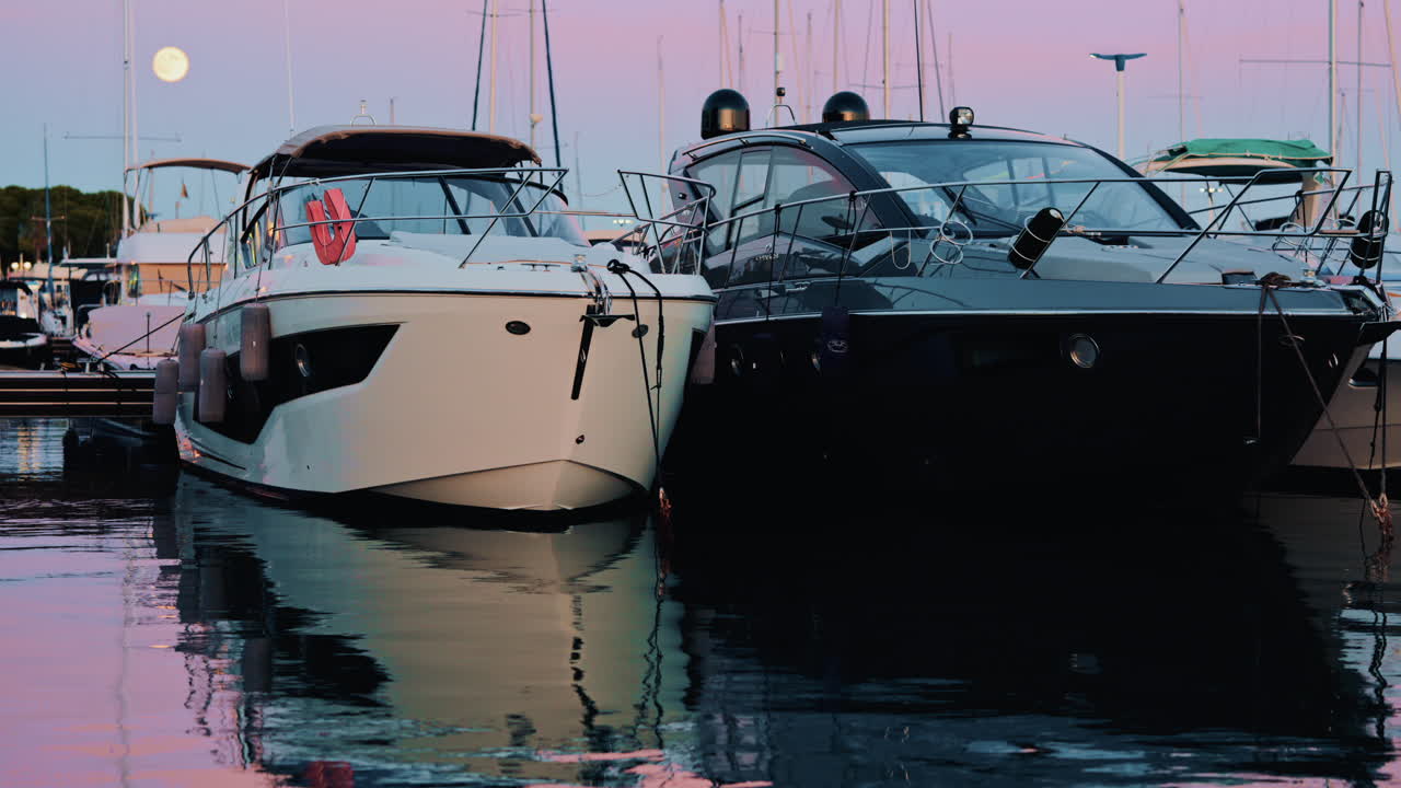 Two modern motorboats moored side by side in a marina just after sunset, under a pink and blue gradient sky with the moon visible