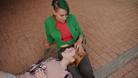 top view of two lesbian girls during their date communicating with each other against the backdrop of paving stones. a blonde girl with a short haircut in a plaid shirt lies on the lap of her girlfriend with a green haircut and a green shirt on her date lgbt couple