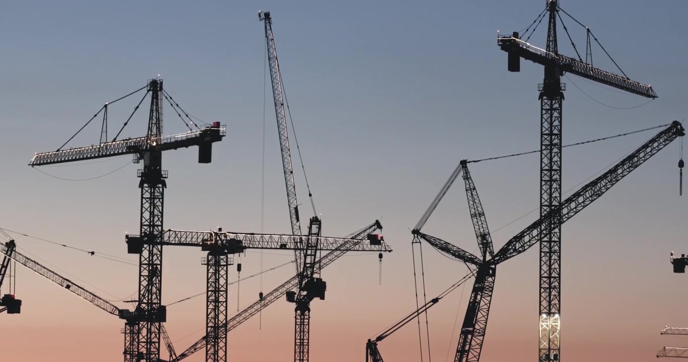 Detailed medium shot of crane silhouettes moving and lifting heavy materials against a colorful evening sky at a technology factory build.