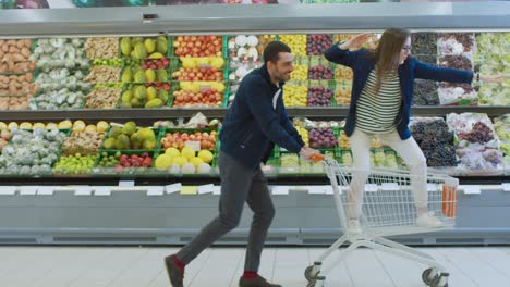 at the supermarket: man pushes shopping cart with woman standing in it, happy couple has fun racing / surfing in a trolley through the fresh produce section of the store. slow motion side view footage.