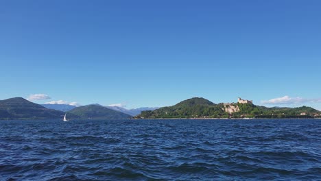 Small-sailboat-sailing-on-Maggiore-lake-agitated-waters-with-mountains-and-Angera-castle-in-background,-Italy