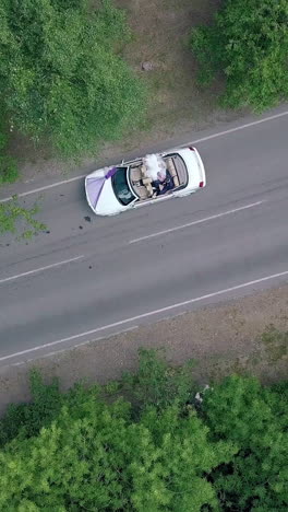 vertical round motion over wedding couple sitting in white celebratory convertible between green trees