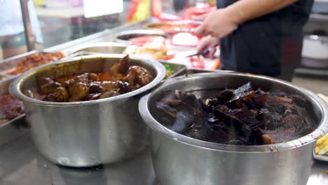 Cook serves traditional Chinese-style stewed pork from large metal pots at a brightly lit Singapore hawker center, with close-up and mid-range shots