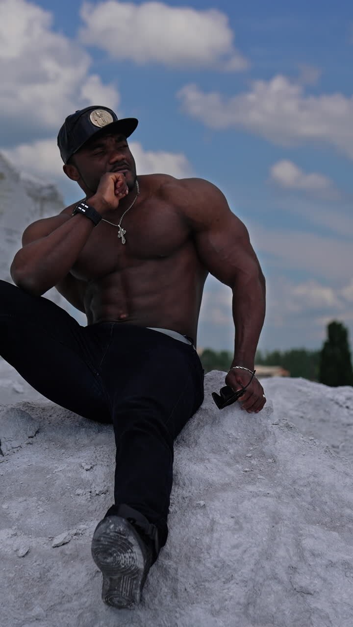 Full length view of the athletic fit model with naked torso sitting at the sand. Strong handsome man posing on blue sky background. Vertical video