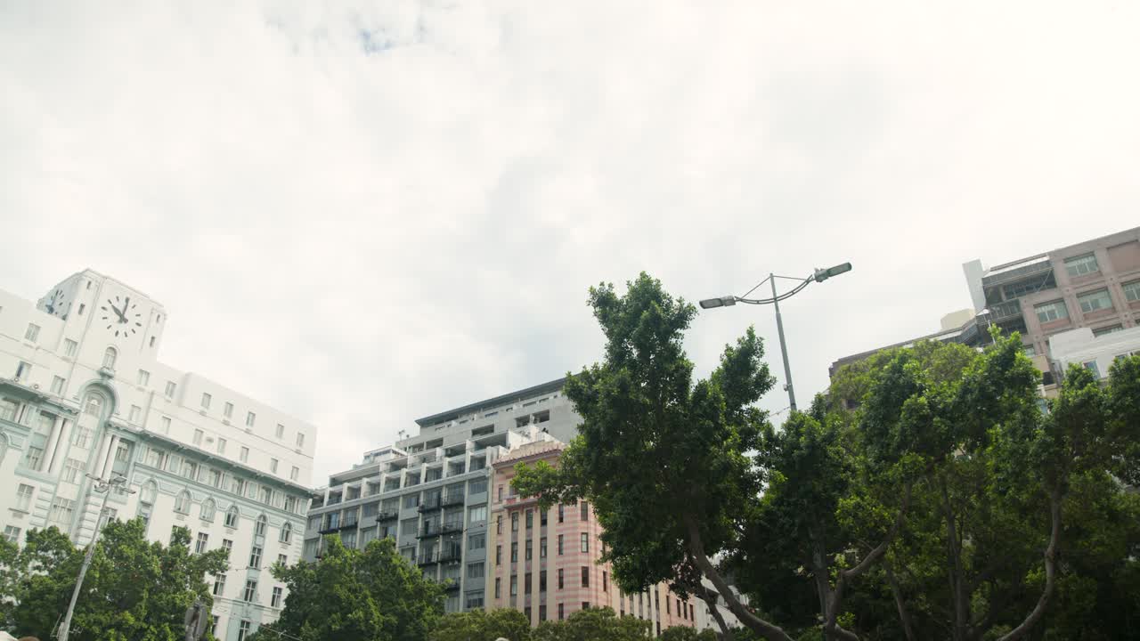 Edifícios, árvore perene emolduram praça urbana enquanto pessoas entram e atravessam para se deslocarem
