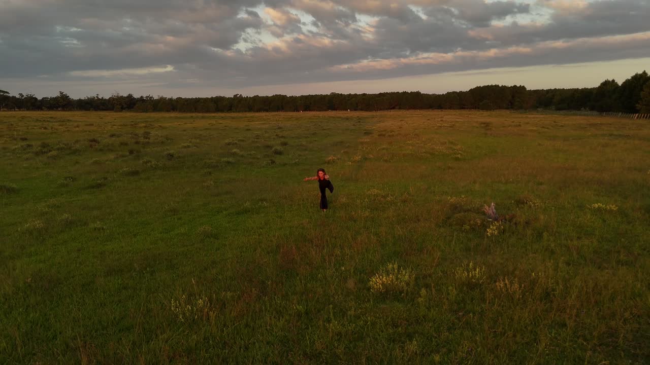 Woman making graceful flow movements, possibly tai chi or dance, in green field at sunset, Punta Del Diablo, Rocha Estate, Uruguay. Aerial drone circling, golden hour