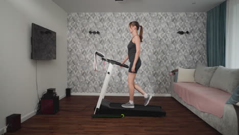 young woman working out on treadmill in stylish living room, walking briskly with arms bent on console, towel draped over handlebars, patterned wall and sofa backdrop