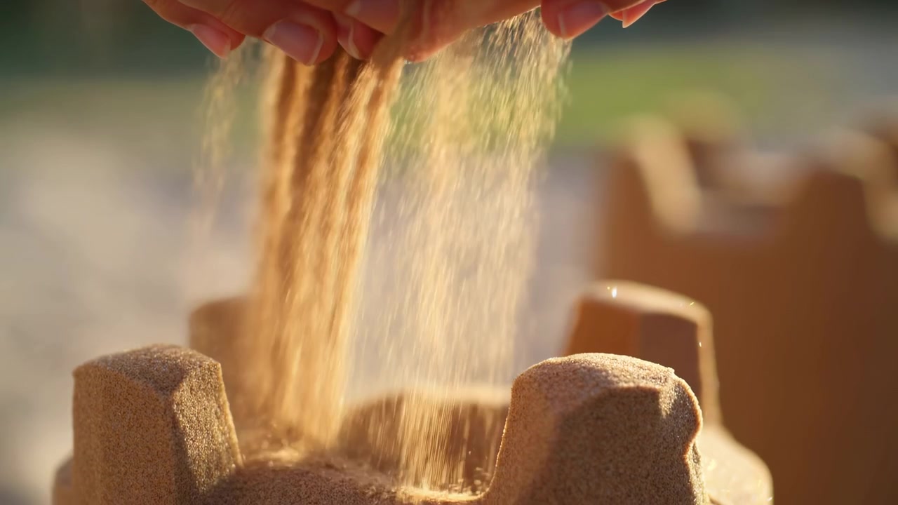 Närbild på två händer som låter en ström av sand rinna genom fingrarna i mjukt gyllene ljus, med en suddig utomhusbakgrund och en lugn naturlig atmosfär.