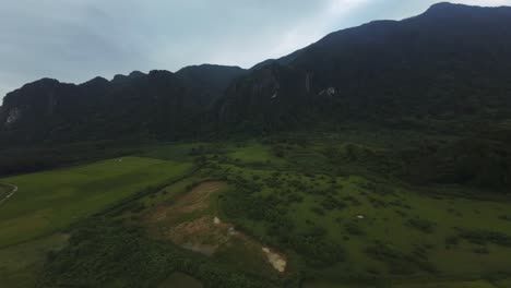 aerial panoramic view over grassy fields near the blue lagoon in vang vieng, laos, capturing lush green fields and mountains under cloudy skies