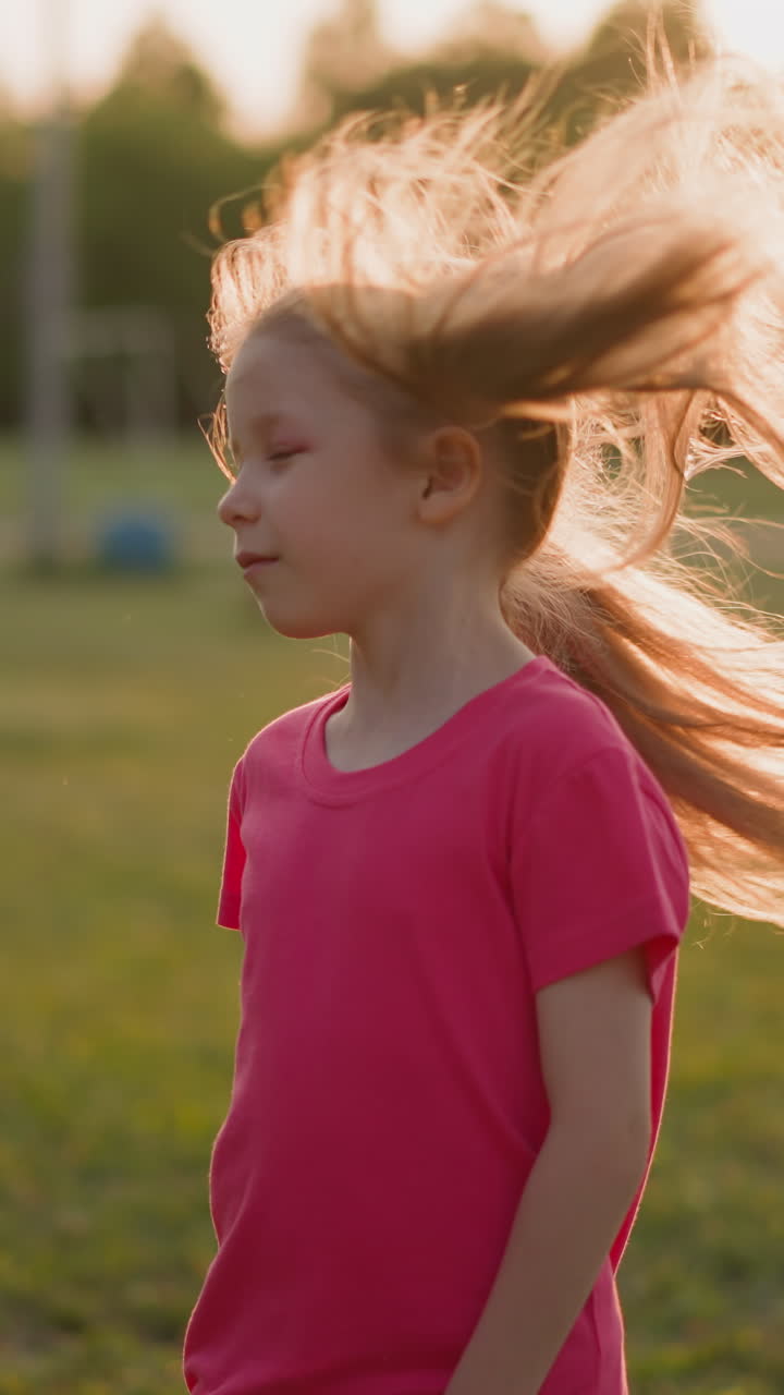 Happy little girl shakes long ponytails on stadium field at back sunset light slow motion. Pretty child in park. Lifestyle and girlhood